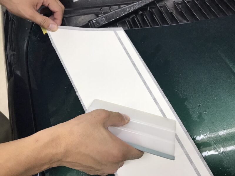 ミラジーノレーシングストライプのインストールマニュアルの施工途中段階のイメージでアプリケーションフィルムの上からスキージでしごいている様子